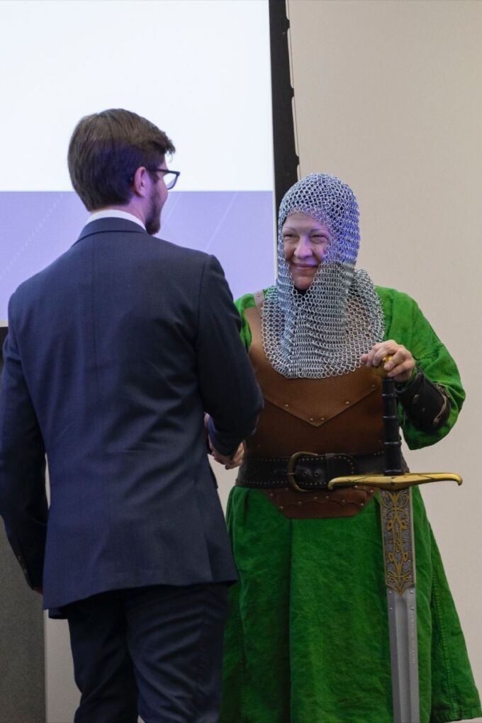 A woman dressed in green dress with a chain mail hood poses with a very large sword while shaking hands with a man in a navy suit