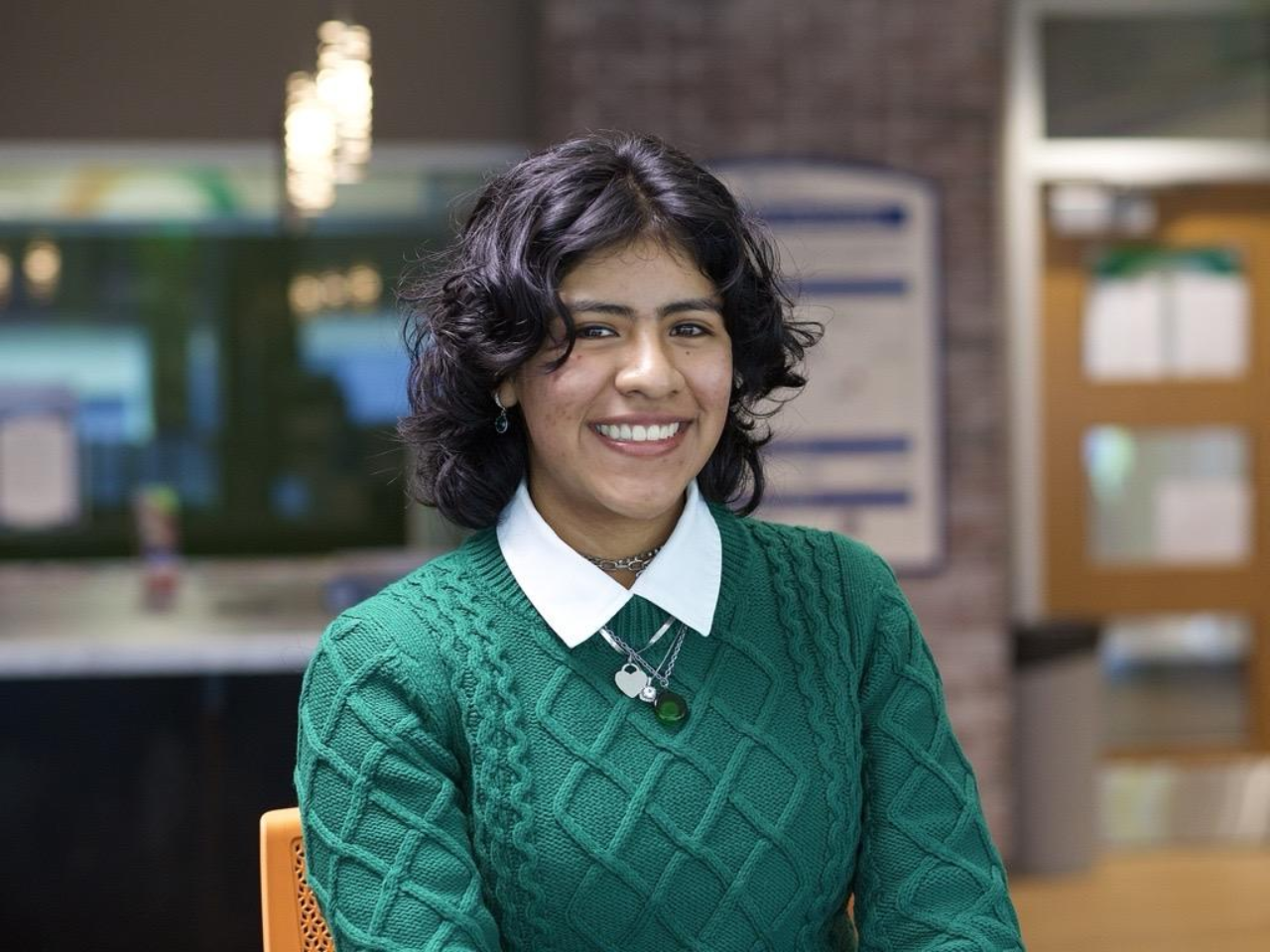 Young student with dark shoulder-length hair smiles broadly in a green cable knit sweater and broad-collared white shirt