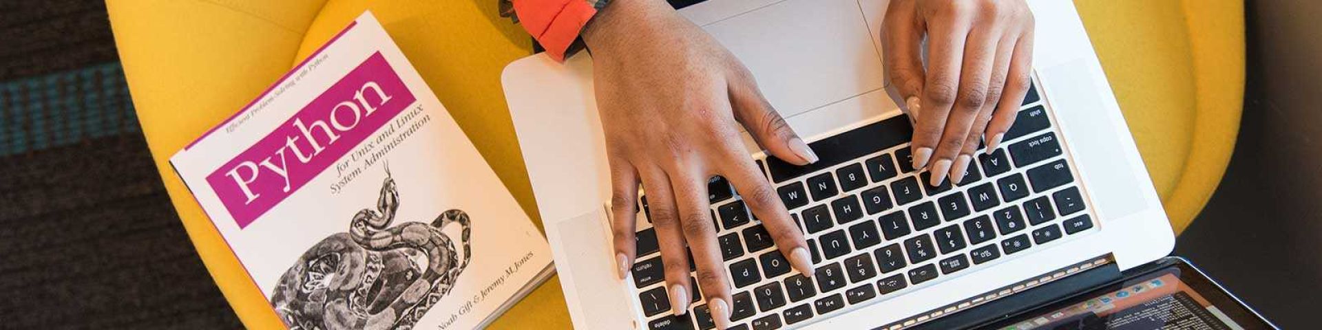 Hands typing on laptop with Python programming book beside