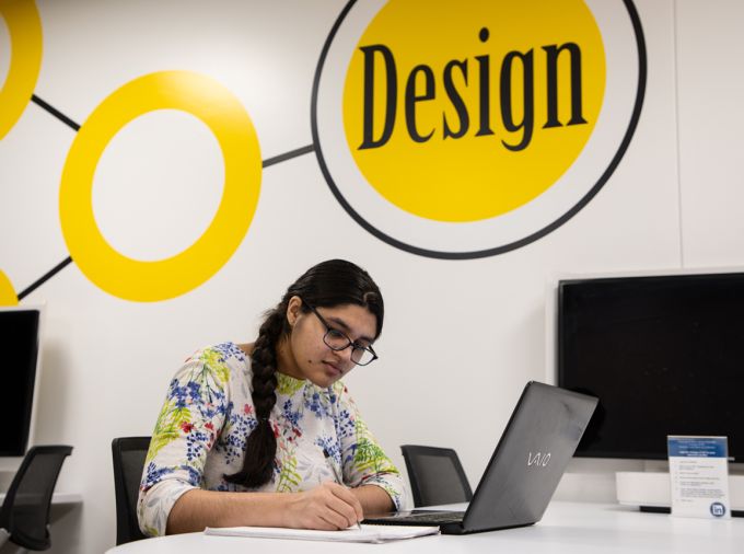 Young woman writing on paper sitting in front of a laptop with a wall behind that says design