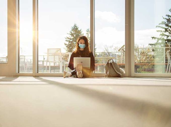Young woman sitting on floor with laptop with glass wall behind and sunshine