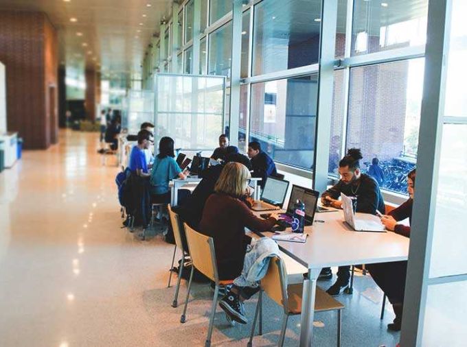 Students in an expansive glass atrium gather at tables and work on laptops