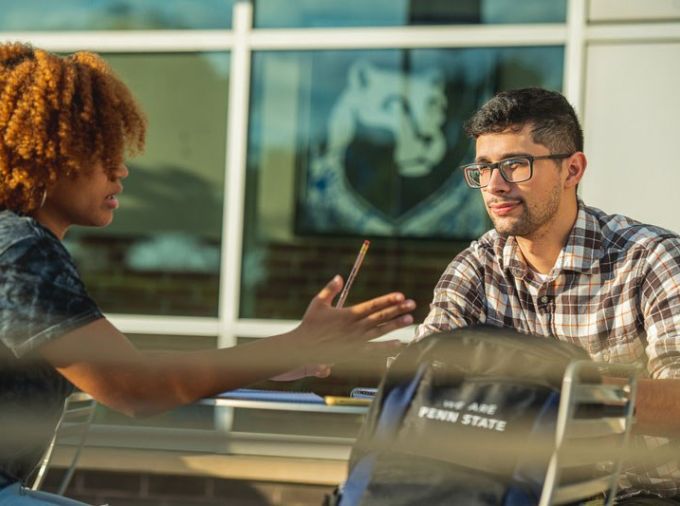Two students sit at a table with laptops having a discussion