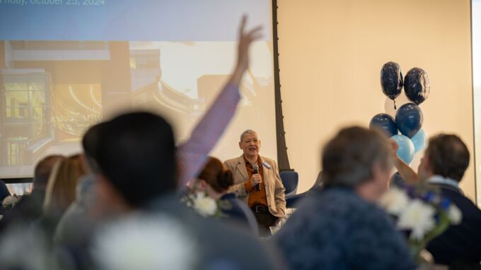 A person speaks to an audience while an individual in the foreground raises their hand.