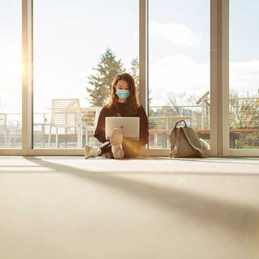 Young woman sitting on floor with laptop with glass wall behind and sunshine