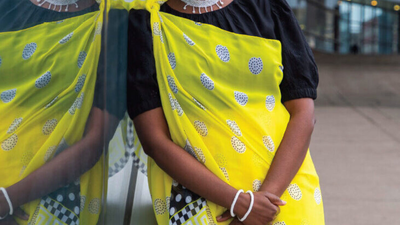 A woman wearing a bright yellow wrap dress poses alongside a window that reveals her reflection