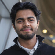 A student with black hair and neatly trimmed beard smiles in a dark zip sweater and white dress shirt