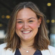 Young woman with shoulder-length brown hair smiles broadly in a navy shirt and white jacket