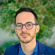 A man with neatly-trimmed hair and thin beard poses outside in front of a wall covered in greenery in round glasses, blue jacket, and blue patterned shirt