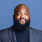 Man with a bald head and beard smiles in a gray jacket and dark shirt in front of a blue background