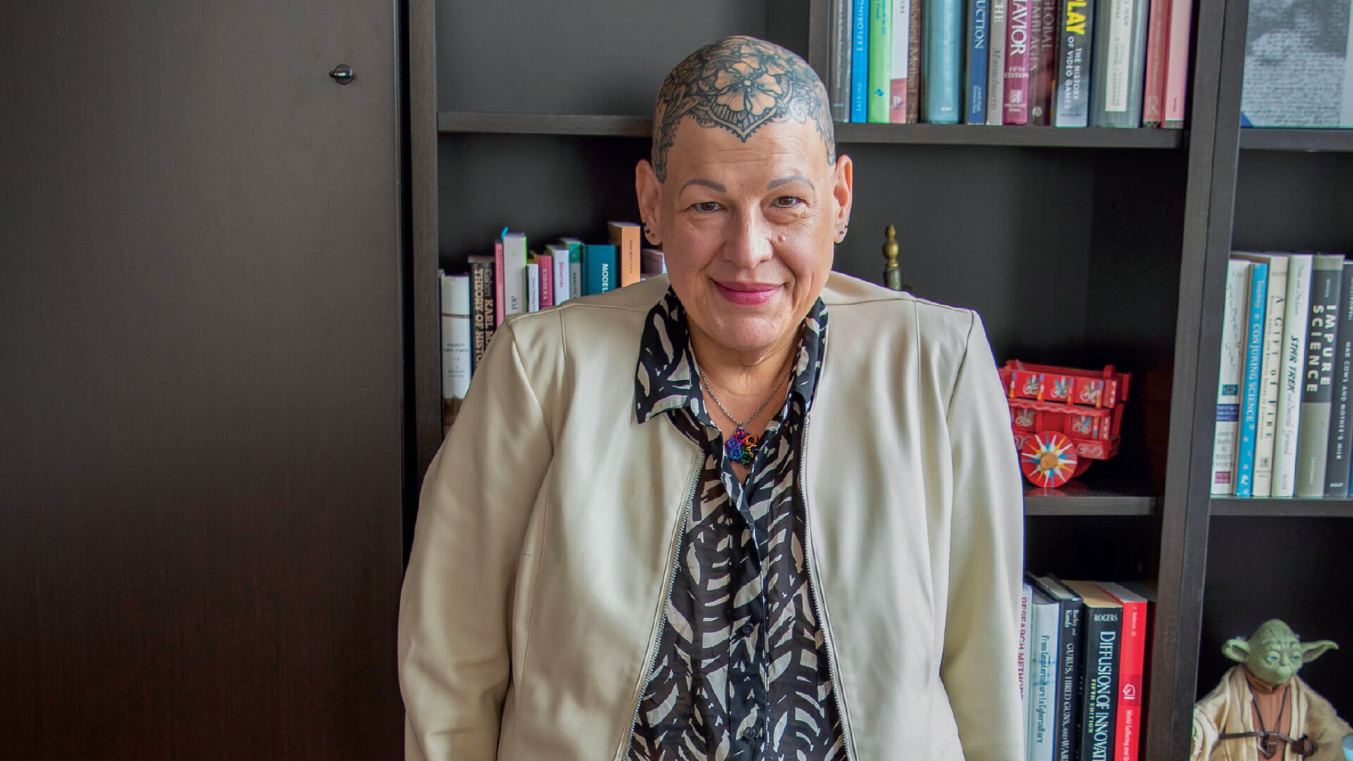 A bald woman with tattooed head poses in an office in front of a bookshelf wearing a tan jacket and patterned shirt