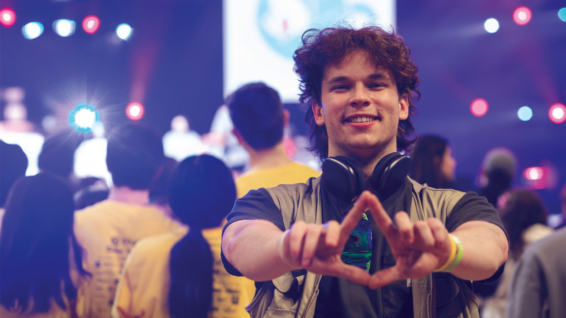 A student forms a diamond shape with his thumbs and index fingers while posing in front of a crowd of students on a brightly lit backdrop in an arena