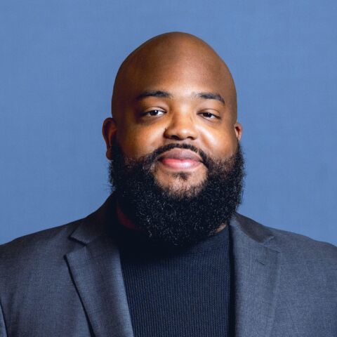 Man with a bald head and beard smiles in a gray jacket and dark shirt in front of a blue background