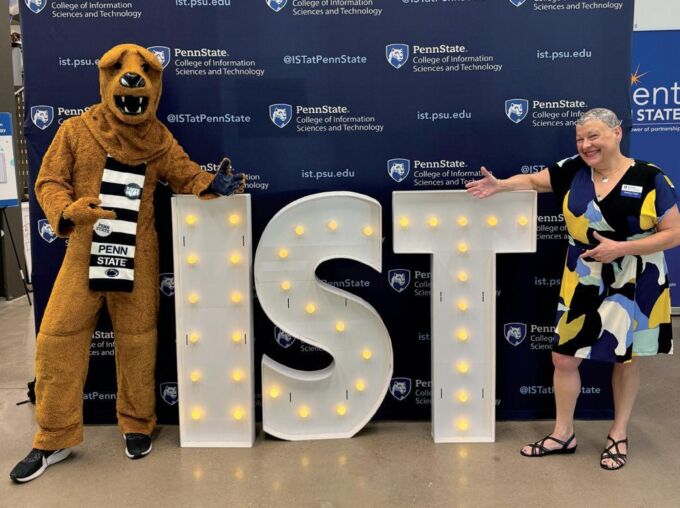 A woman in a bright patterned dress with bald, tattooed head poses in front of a lighted IST sign with a mountain lion sports mascot dressed with navy blue and white stripped scarf