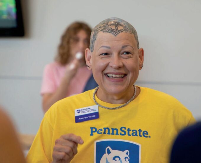 A bald woman with a tattooed head smiles in a yellow tee shirt printed with the Penn State logo