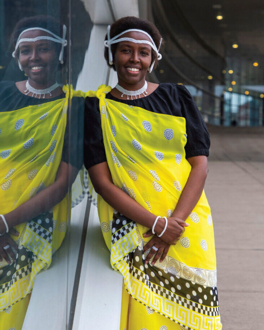 A woman wearing a bright yellow wrap dress poses alongside a window that reveals her reflection